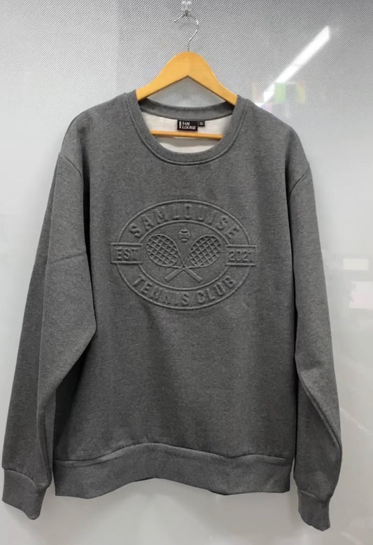 The SamLouise Tennis Club Embossed Sweatshirt, draped on a hanger, displays an emblem of crossed rackets and SamLouise Tennis Club EST 2021 on the front. This limited edition merges timeless style with a modern flair, ideal for tennis fans and trendsetters alike.