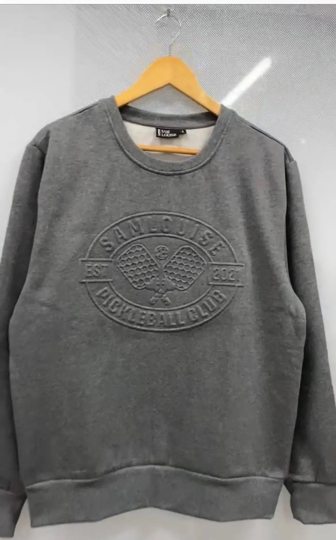 The SamLouise Pickleball Club Embossed Sweatshirt is a gray crewneck limited edition style with an embossed logo and paddle design, shown on a wooden hanger against a light background.