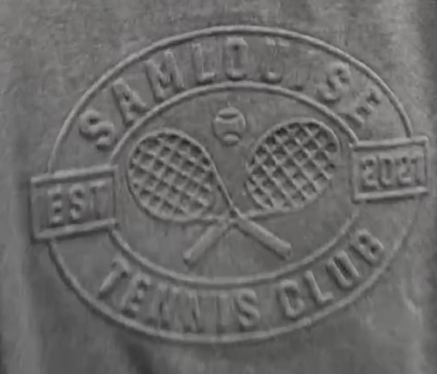 The SamLouise Tennis Club Embossed Sweatshirt, draped on a hanger, displays an emblem of crossed rackets and SamLouise Tennis Club EST 2021 on the front. This limited edition merges timeless style with a modern flair, ideal for tennis fans and trendsetters alike.