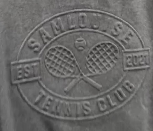 The SamLouise Tennis Club Embossed Sweatshirt, draped on a hanger, displays an emblem of crossed rackets and SamLouise Tennis Club EST 2021 on the front. This limited edition merges timeless style with a modern flair, ideal for tennis fans and trendsetters alike.