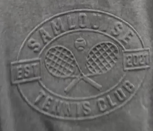The SamLouise Tennis Club Embossed Sweatshirt, draped on a hanger, displays an emblem of crossed rackets and SamLouise Tennis Club EST 2021 on the front. This limited edition merges timeless style with a modern flair, ideal for tennis fans and trendsetters alike.