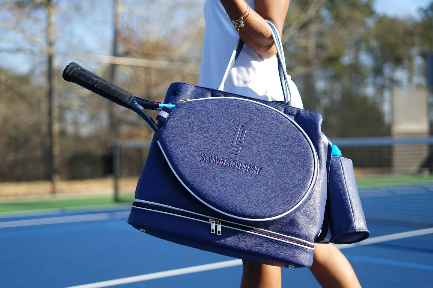 The SamLouise London 77 Tennis Tote from SamLouise is a chic sports tote bag in blue with white accents and a detachable strap. It includes a zippered bottom compartment, an insulated bottle holder, and a cylindrical pouch on the side.